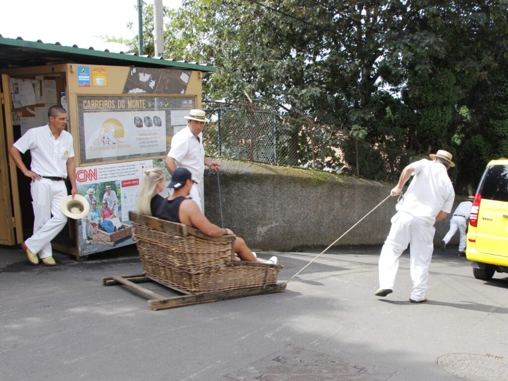 Zwei weiss gekleidete Männer ziehen einen Korbschlitten, in welchem zwei Personen sitzen, eine Strasse hinunter