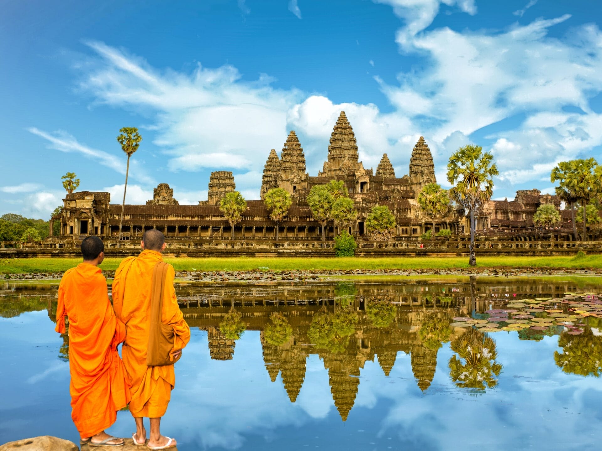Zwei orange gekleidete Mönche stehen am Wasser, in dem sich die Tempelanlage von Ankor Wat und der blaue Himmel spiegelt.