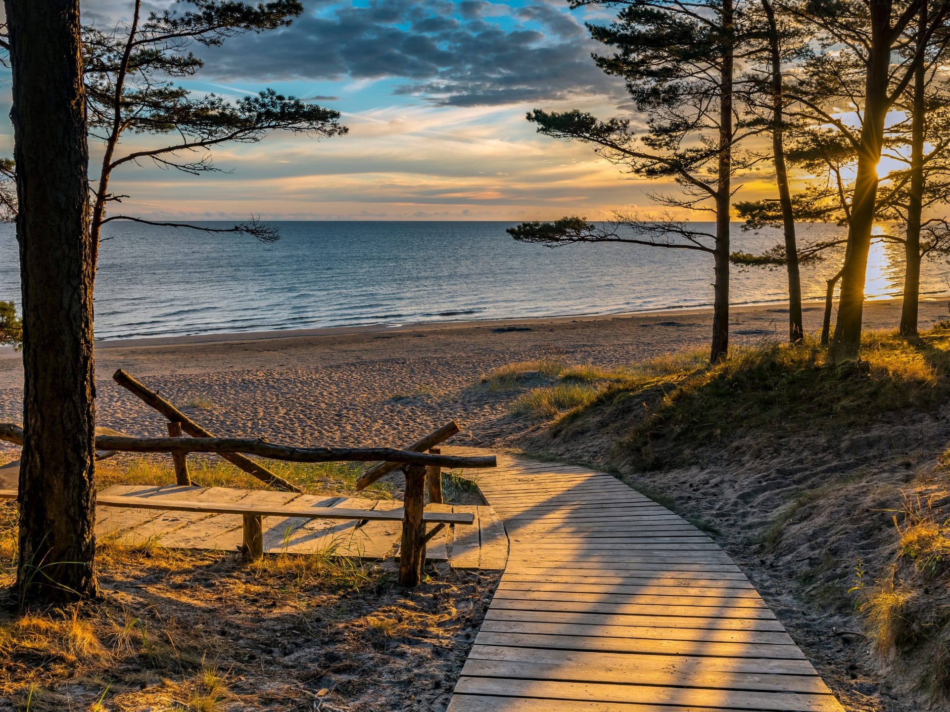 Holzsteg am Strand von Jurmala in Lettland flankiert von Bäumen im goldenen Abendlicht. Im Hintergrund ist das Meer zu sehen.