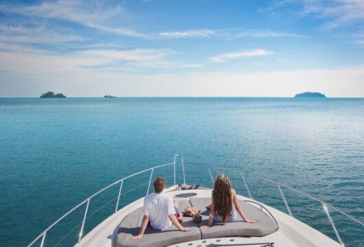 Eine Frau und ein Mann sitzen auf dem Bug eines Schiffes, das auf dem Meer fährt. Am Horizont sind Inseln zu sehen.