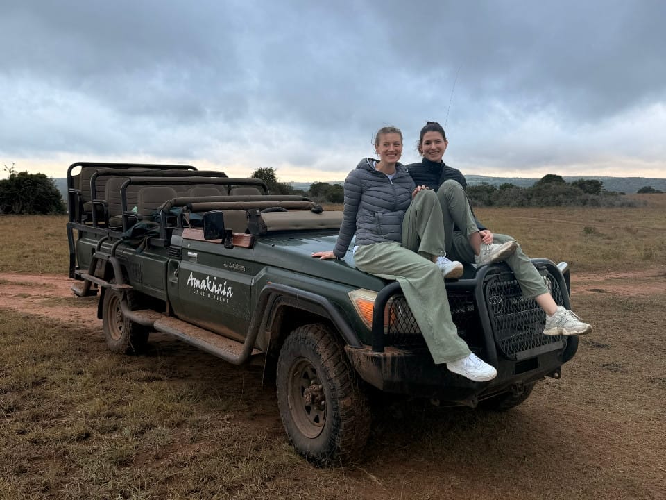 Zwei Vögele-Mitarbeiterinnen sitzen bei bewölktem Himmel auf dem Safari-Fahrzeug in der Steppe des Amakhala Game Reserves in Südafrika.