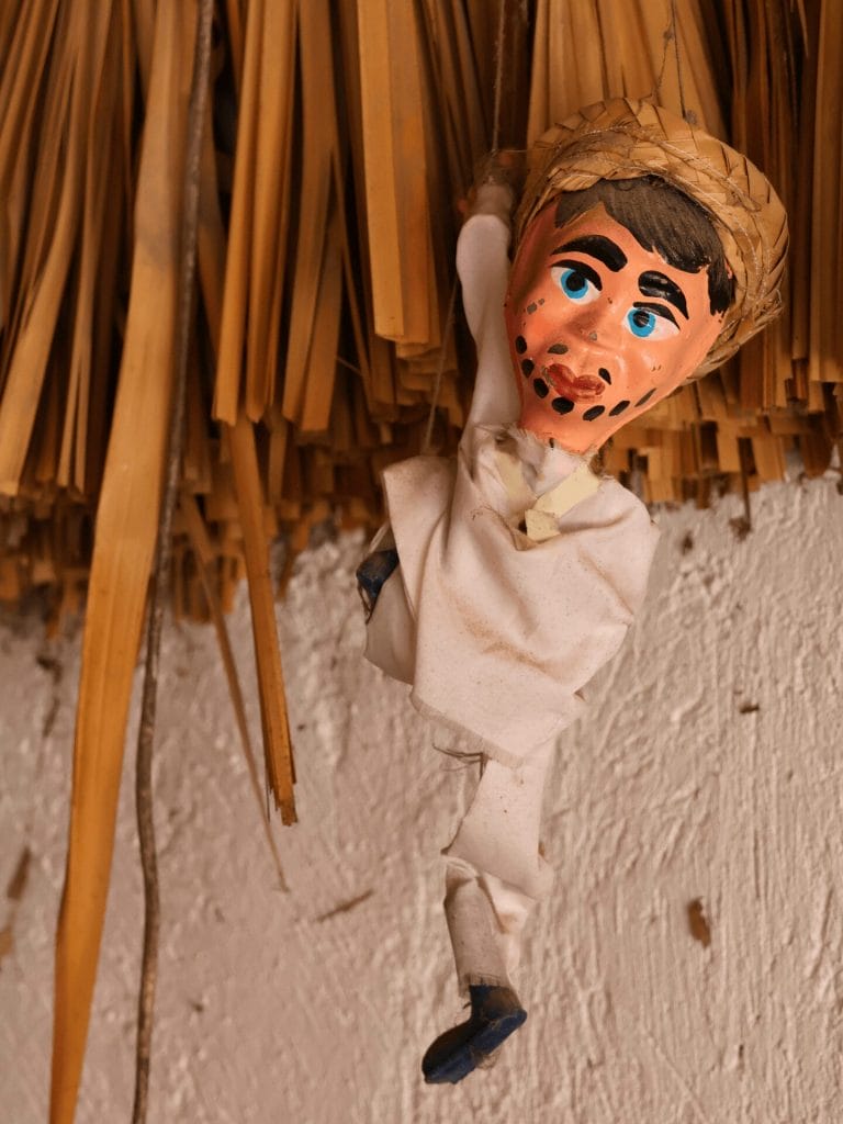 Eine kleine handgemachte Puppe mit weissen Stoffkleidern, schwarzen Schuhen und einem Strohhut hängt an einer Strohhalm-Decke.