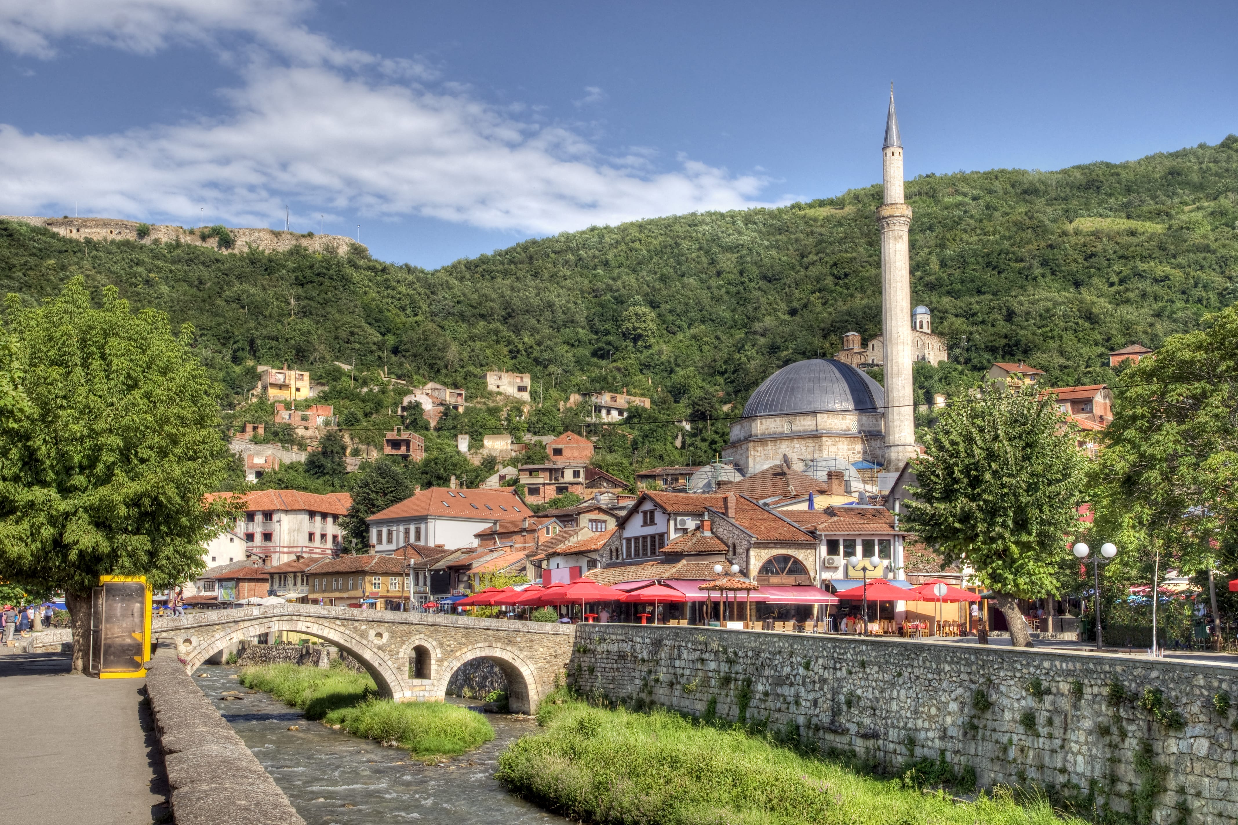 Die historische Altstadt von Prizren mit Moschee und Minarett bei blauem Himmel, eingebettet in grüne Hügelzüge. Im Vordergrund fliesst ein kanalisierter Fluss, über den eine Bogenbrücke führt.