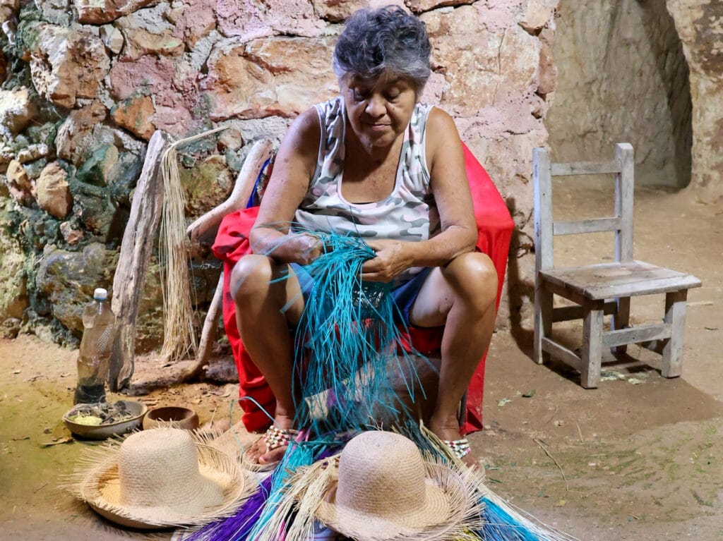 Eine mexikanische Frau mit schwarzen, graumelierten Haaren, sitzt in einer Kellerhöhle auf einem Stuhl beim Flechten eines türkisfarbenen Huts. Am Boden vor ihren Füssen liegen zwei fast fertig geflochtene, naturfarbene Strohhüte sowie farbige Strohhalme.
