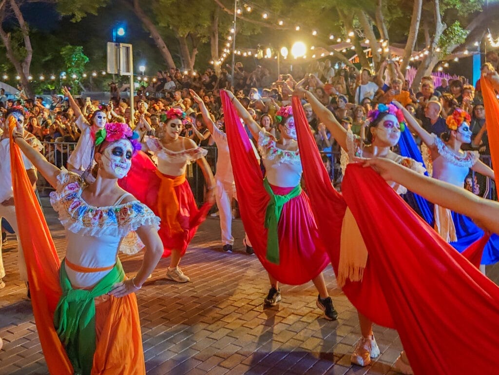 Traditionelle Tänzerinnen mit kunstvollem Catrina-Make-up und bunten Gewändern beim ausgelassenen Tanz während der Feierlichkeiten zum Día de los Muertos in Mexiko vor begeistertem Publikum