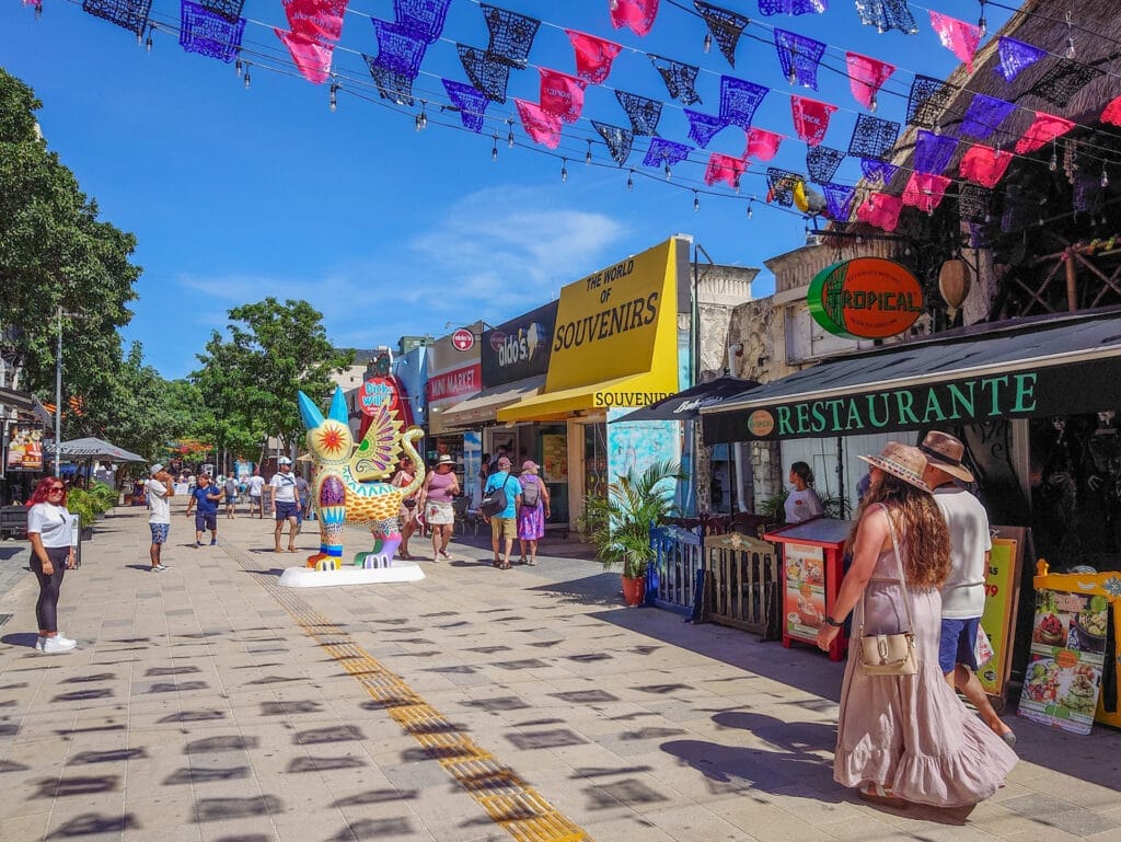 Gäste flanieren bei sonnigem Wetter durch eine belebte Einkaufsstrasse in Playa del Carmen, Mexiko, geschmückt mit bunten Wimpelketten und einer grossen Alebrije-Fantasiefigur.