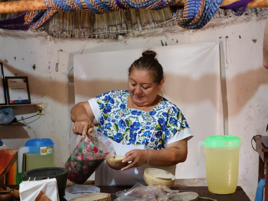 Einheimische Mexikanerin in traditioneller, blau bestickter Kleidung schenkt ein lokales Getränk aus einem Krug in eine Naturschale ein.