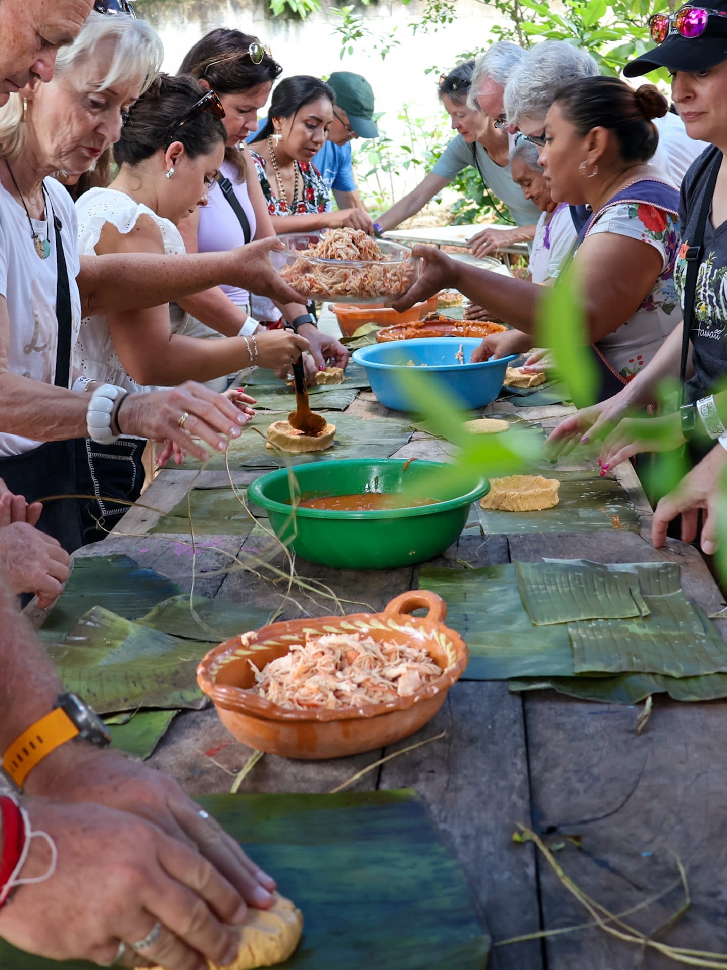 Gäste und Einheimische beim gemeinsamen Zubereiten traditioneller Speisen an einem langen Holztisch, belegt mit Bananenblättern und Schalen voller Zutaten