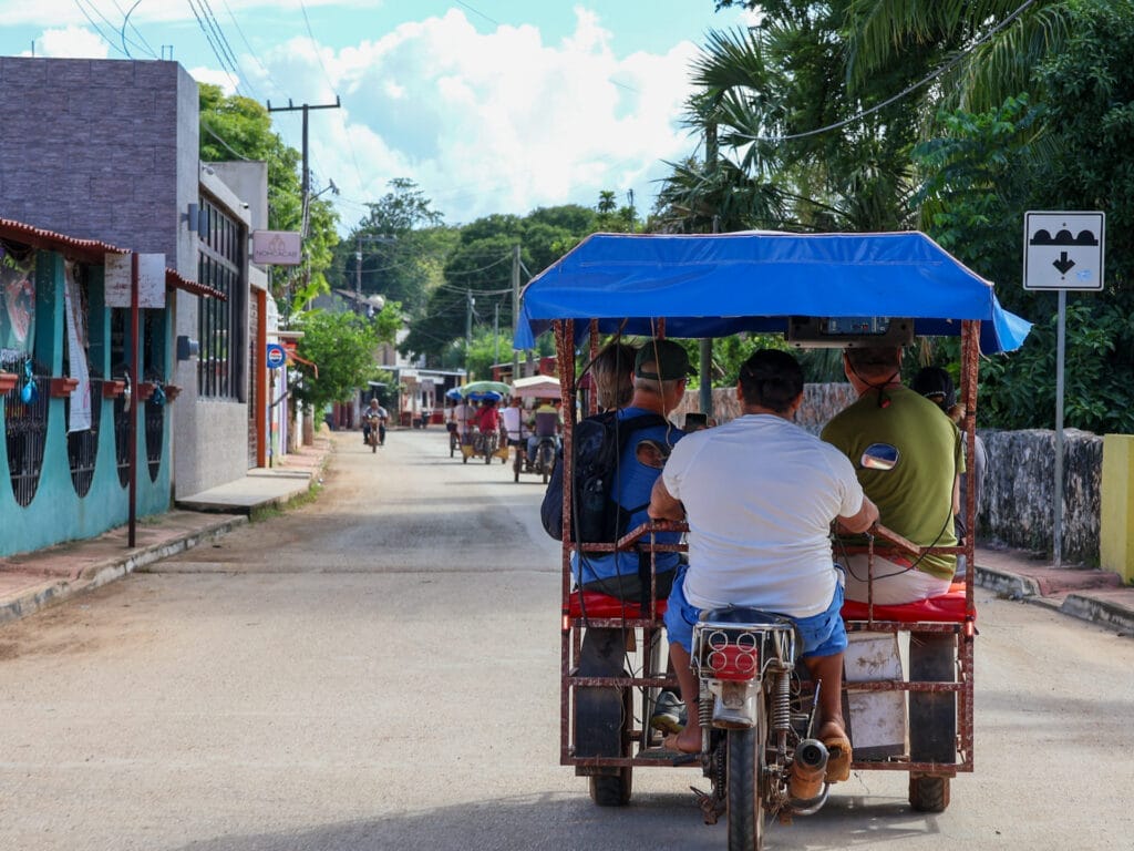 Nahaufnahme eines blauen Mototaxis mit Gästen auf einer Dorfstrasse in Mexiko, flankiert von bunten Häusern und einer Steinmauer