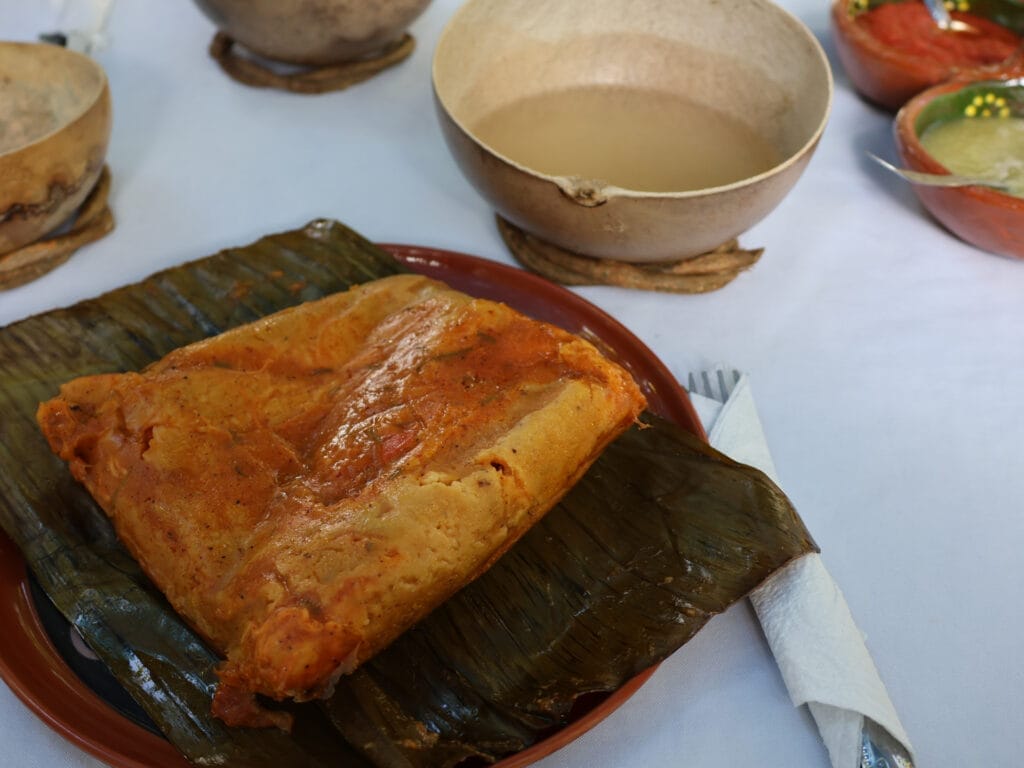 Traditionelles mexikanisches Mukbilpollo Tamale, im Bananenblatt gegart und serviert, auf einem Teller vor typischen Schalen auf einem weissen Tischtuch.