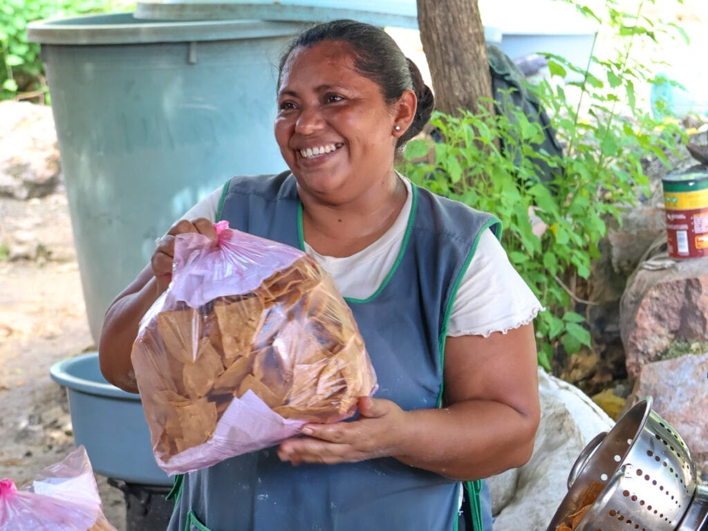 Lächelnde Einheimische in Schürze präsentiert stolz eine grosse Tüte mit frischen, selbstgemachten Tortilla-Chips in einer mexikanischen Aussenküche