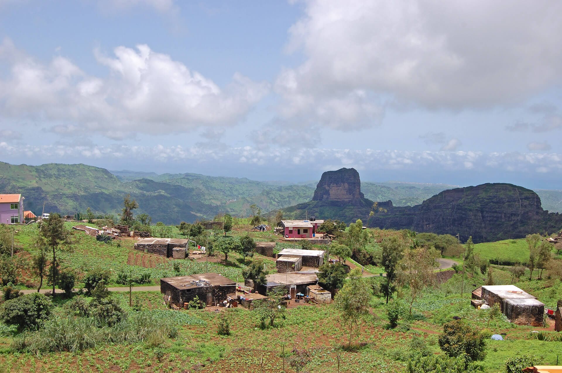 Panoramablick über die üppig grüne Berglandschaft der Kapverden. Im Vordergrund liegt ein kleines Dorf mit einfachen Häusern an einem Hang, dahinter ragt ein markanter Tafelberg auf.