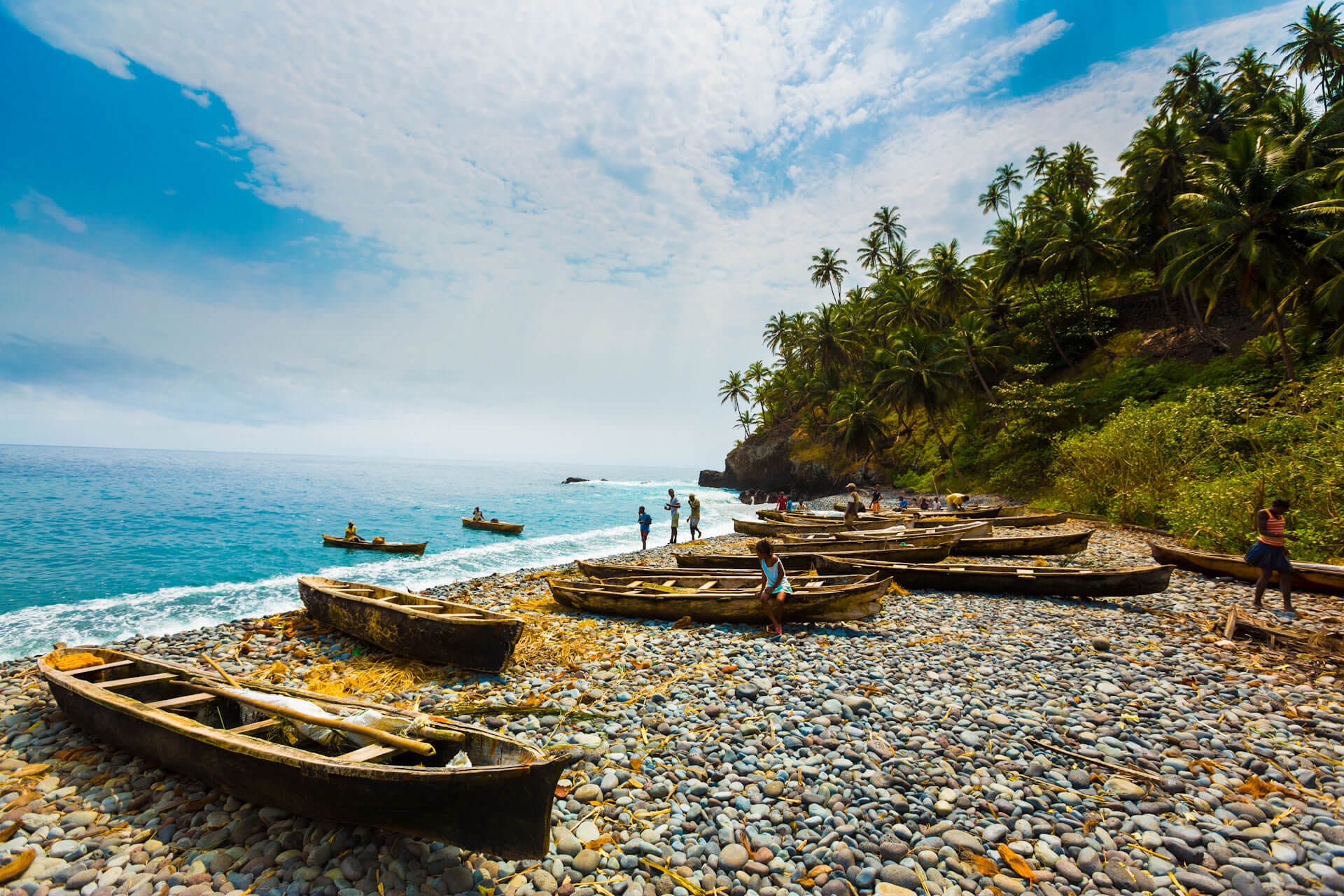Eine malerische Szene an einem tropischen Kieselstrand, wo zahlreiche traditionelle Holzkanus am Ufer liegen. Im Hintergrund säumen hohe Palmen die Küste unter einem strahlend blauen Himmel mit weissen Wolken, während das türkisfarbene Meer sanft an den Strand rollt.