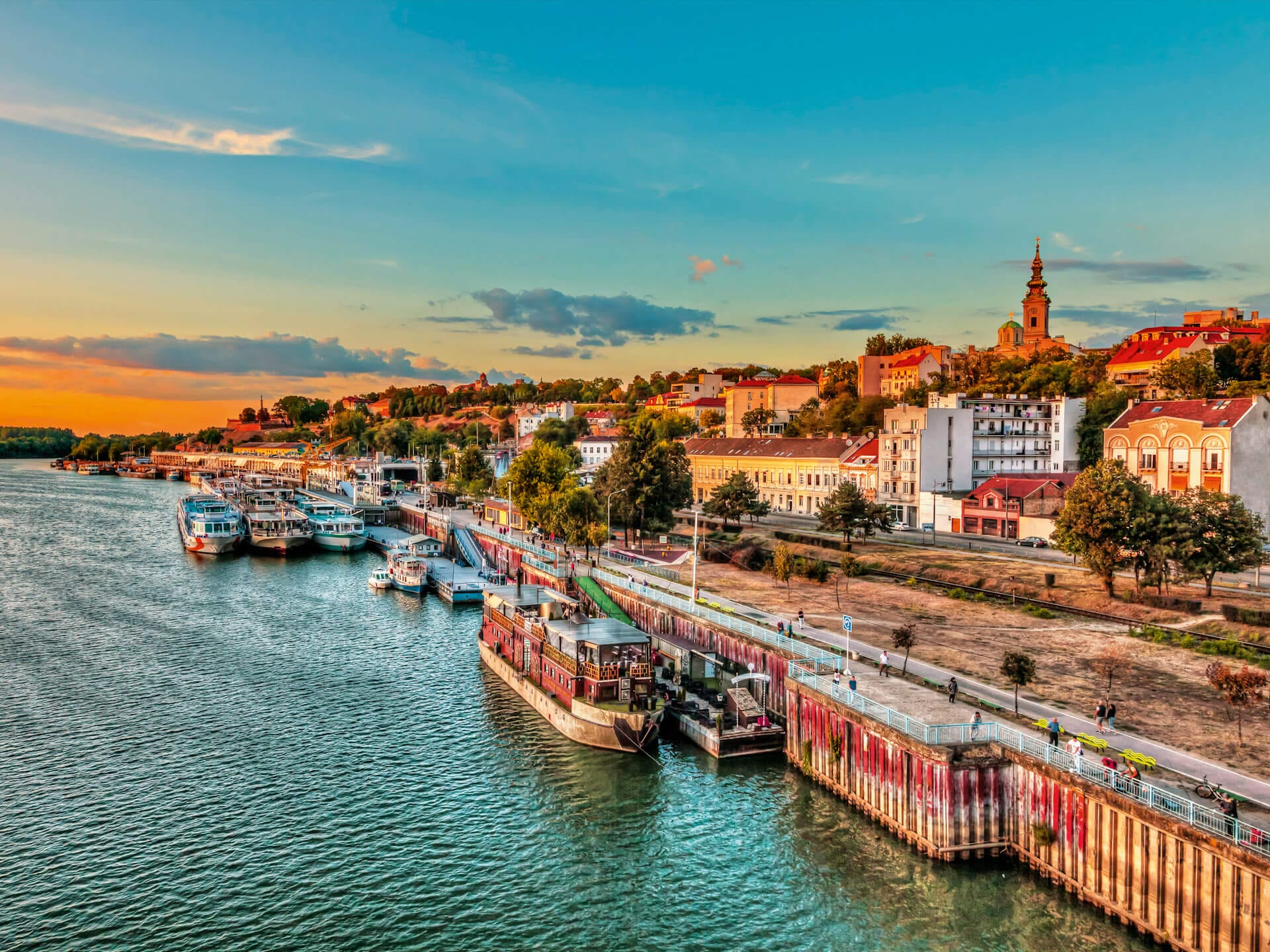 Am Donau-Ufer der serbischen Hauptstadt Belgrad legen die Flussschiffe an. Am Himmel ist eine malerische Abendstimmung zu sehen. Entlang der Uferpromenade spazieren Personen.