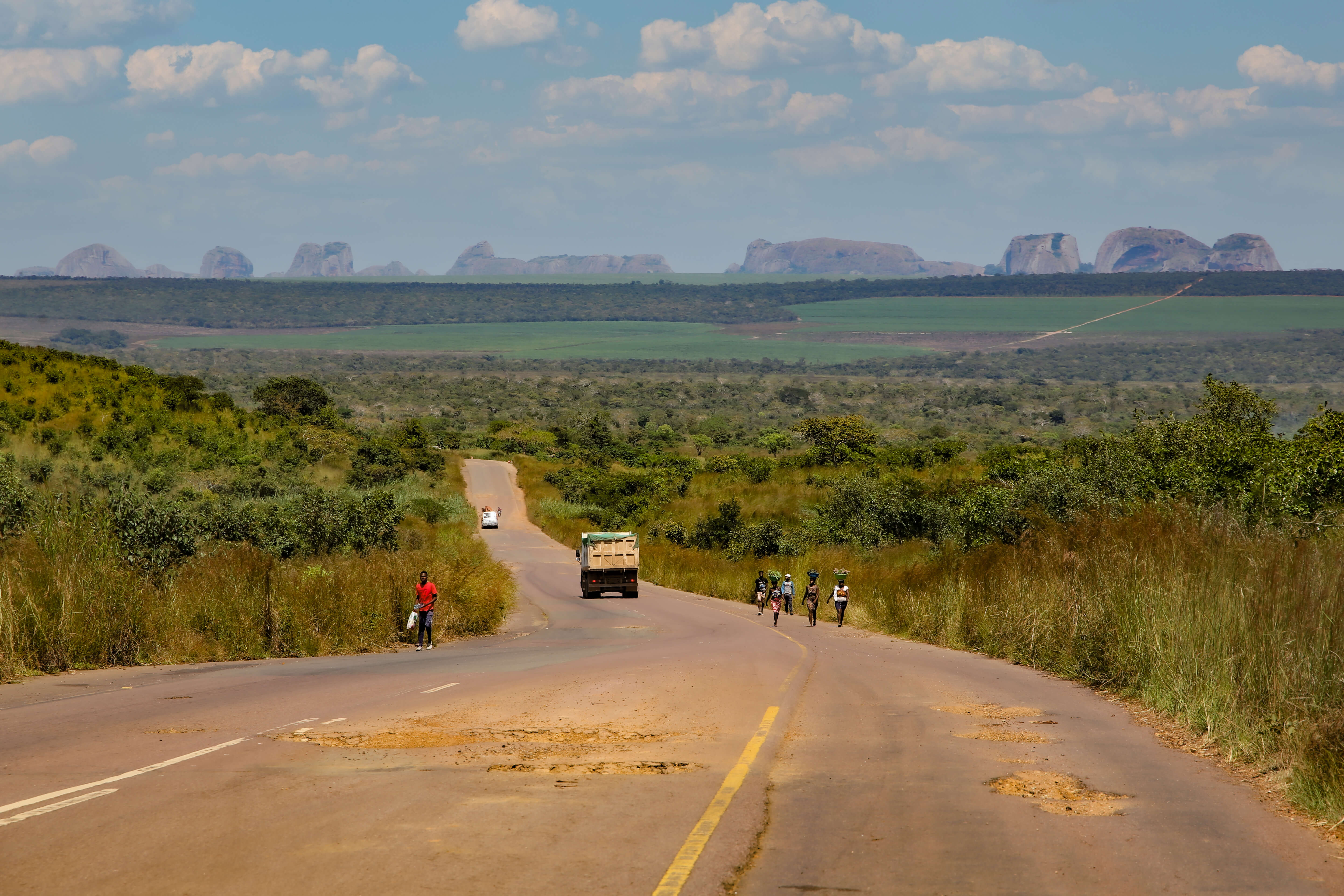 Ländliche Landschaft in Angola mit einer geschwungenen Strasse, auf der ein Lastwagen, ein Kleinwagen und Personen unterwegs sind. Im Hintergrund erstrecken sich grüne Felder und markante Felsformationen unter einem bewölkten Himmel.
